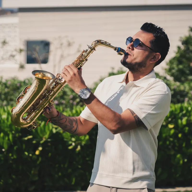 Manu playing alto saxophone outdoors in sunglasses and white shirt