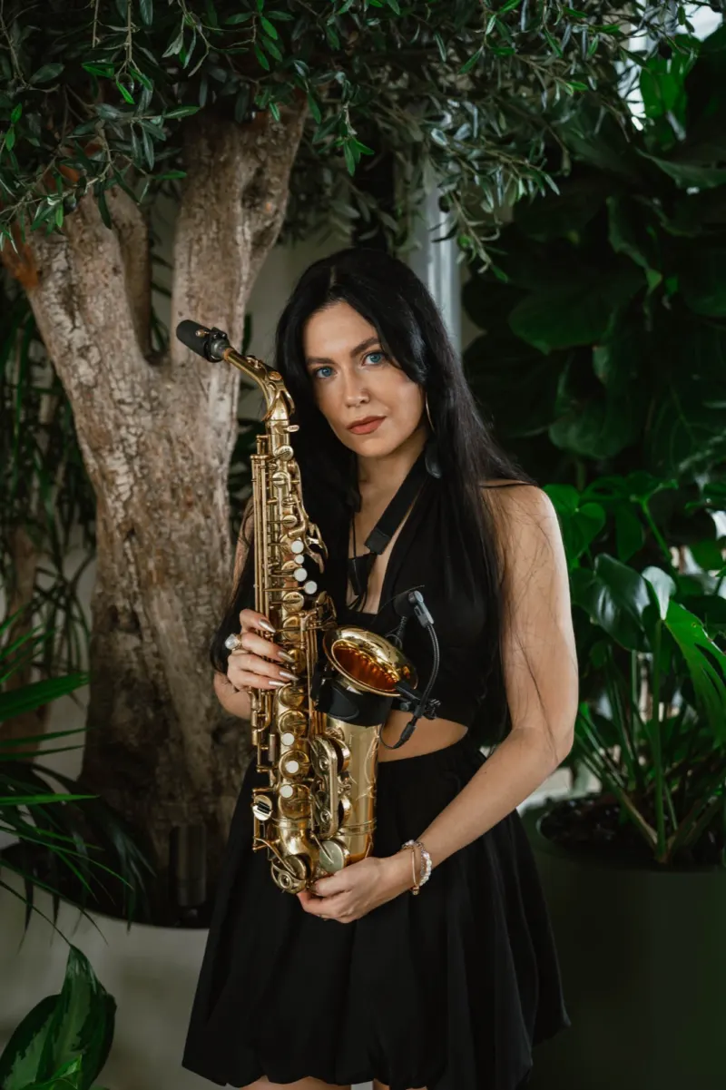 Katrin playing saxophone in a black dress against deep foliage
